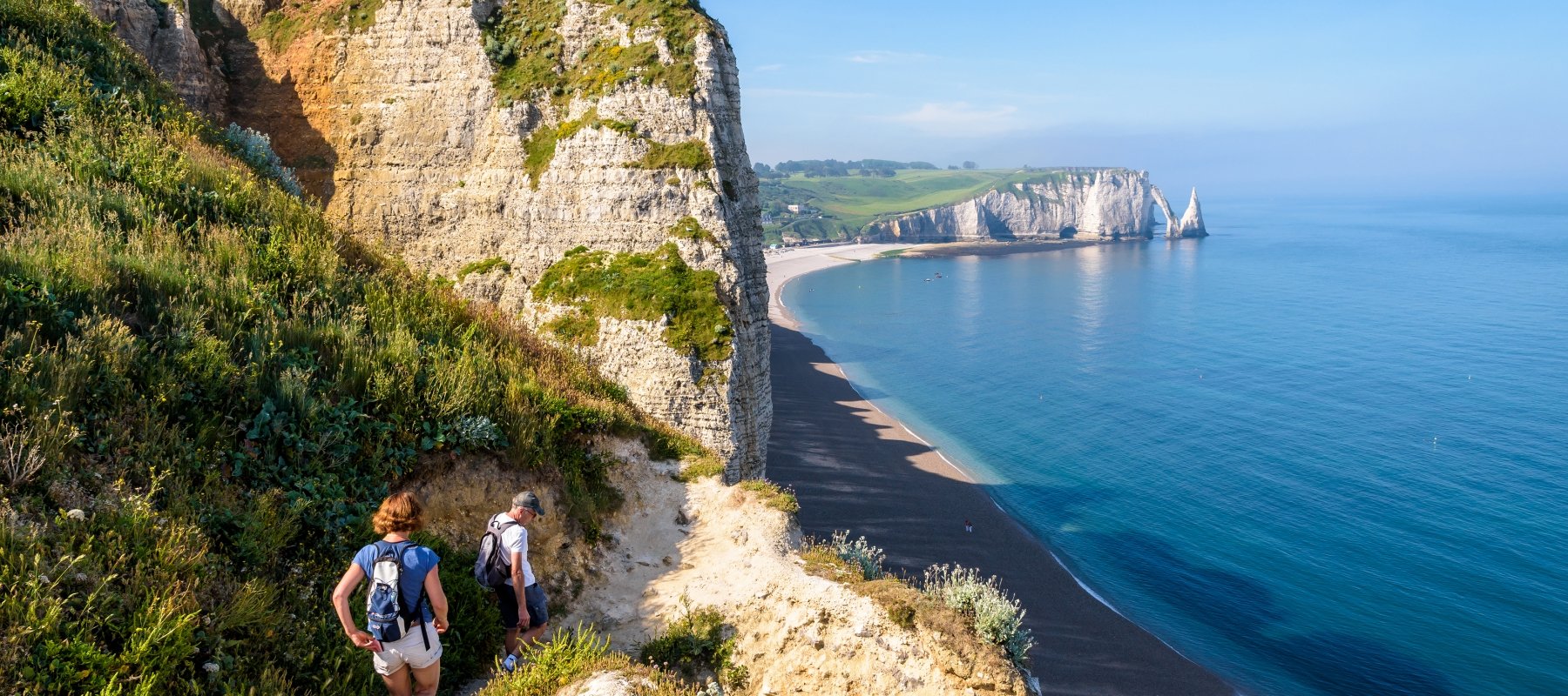A couple in Normandy, France, walking down the rock stairs to the beach at the foot of the Amont cliff. In the distance the Aval cliff, the arch and the Needle.